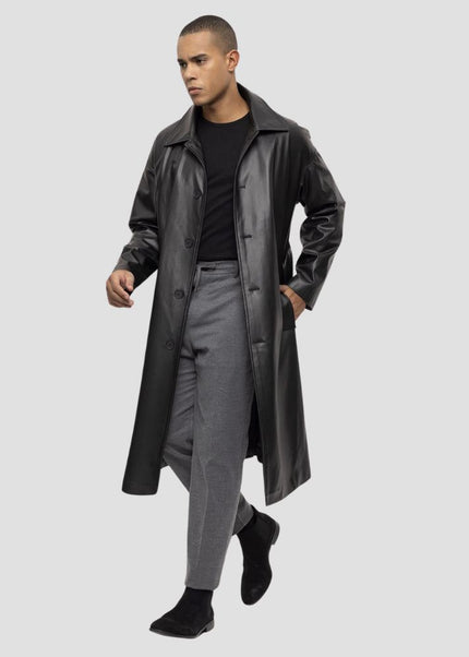 Man wearing a long black coat over a black shirt and gray pants on a light gray background