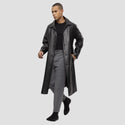 Man wearing a long black coat over a black shirt and gray pants on a light gray background