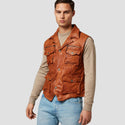 Man wearing a brown leather vest over a beige long-sleeve shirt and blue jeans on a light gray background